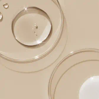 Clear skincare serum droplets in glass petri dishes on a beige background